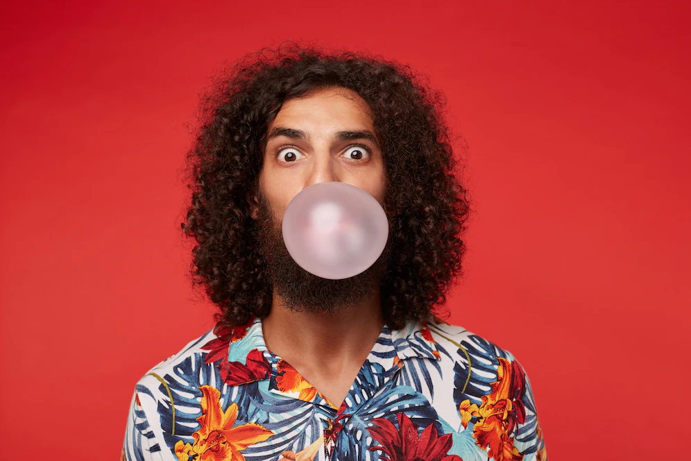 astonished-attractive-young-brunette-curly-man-with-beard-wearing-multi-colored-flowered-shirt-while-posing-rounding-eyes-inflating-bubble-with-pink-gum_295783-5765