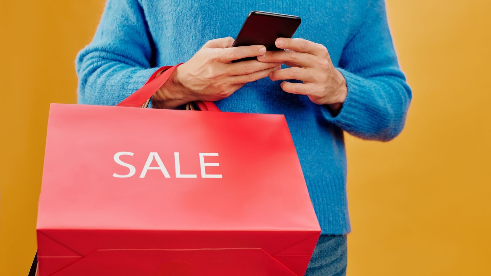 A shopper holding a smartphone and a red bag printed with the word sale