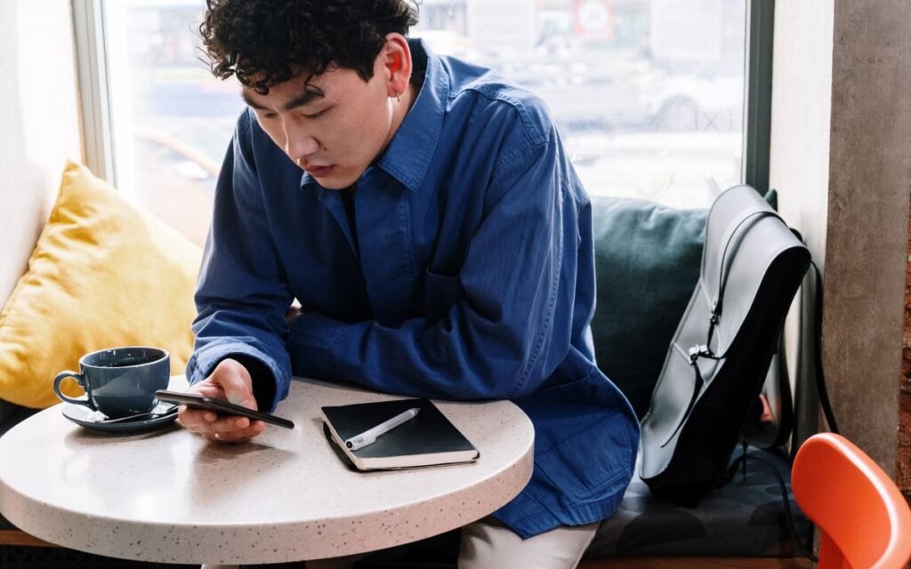 young man checking his phone at a cafe table with a coffee and notebook