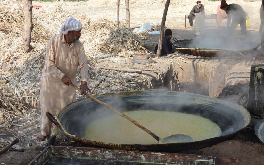 Man in a white turban stirring a huge pan of boiling sugarcane juice outdoors