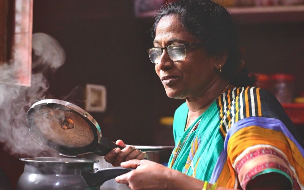 Indian woman in glasses lifting the lid of a steaming pressure cooker in her kitchen