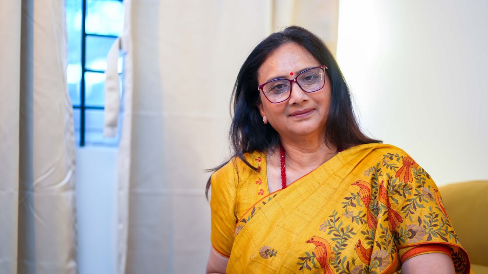 Indian woman in her fifties wearing a yellow saree and glasses, sitting indoors