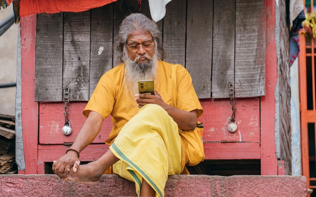 older Indian man in a yellow kurta reading his smartphone on a doorstep