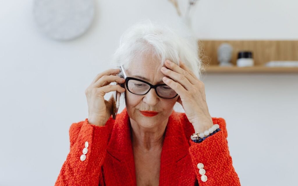 Older woman in glasses holding her head while on a phone call