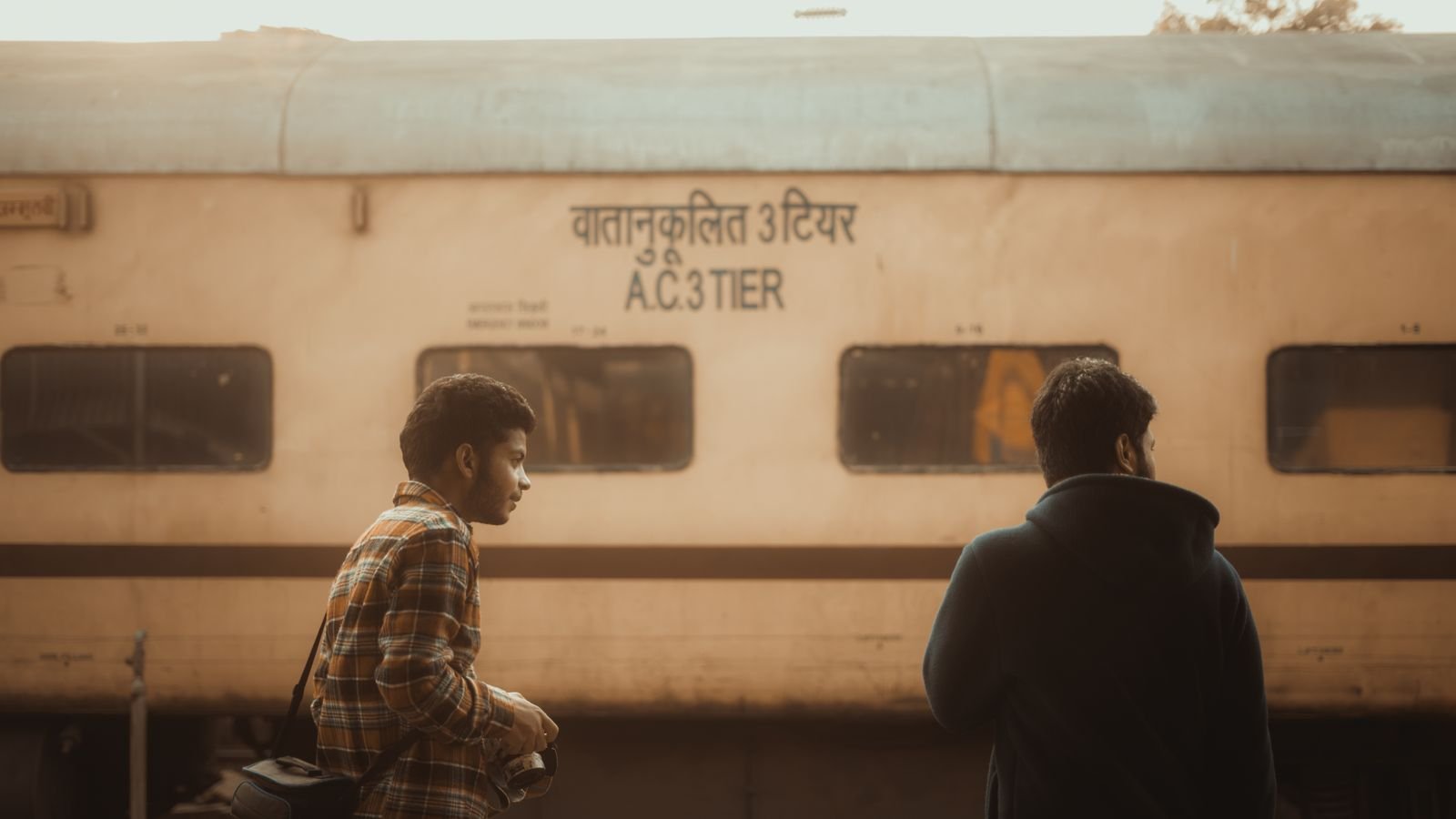 Two passengers standing beside an AC 3-tier train coach at an Indian station